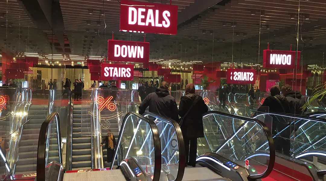 Rote Leuchtschilder an Rolltreppen weisen auf "Deals downstairs" hin - eine klare POS Kommunikation zur Verkaufsförderung